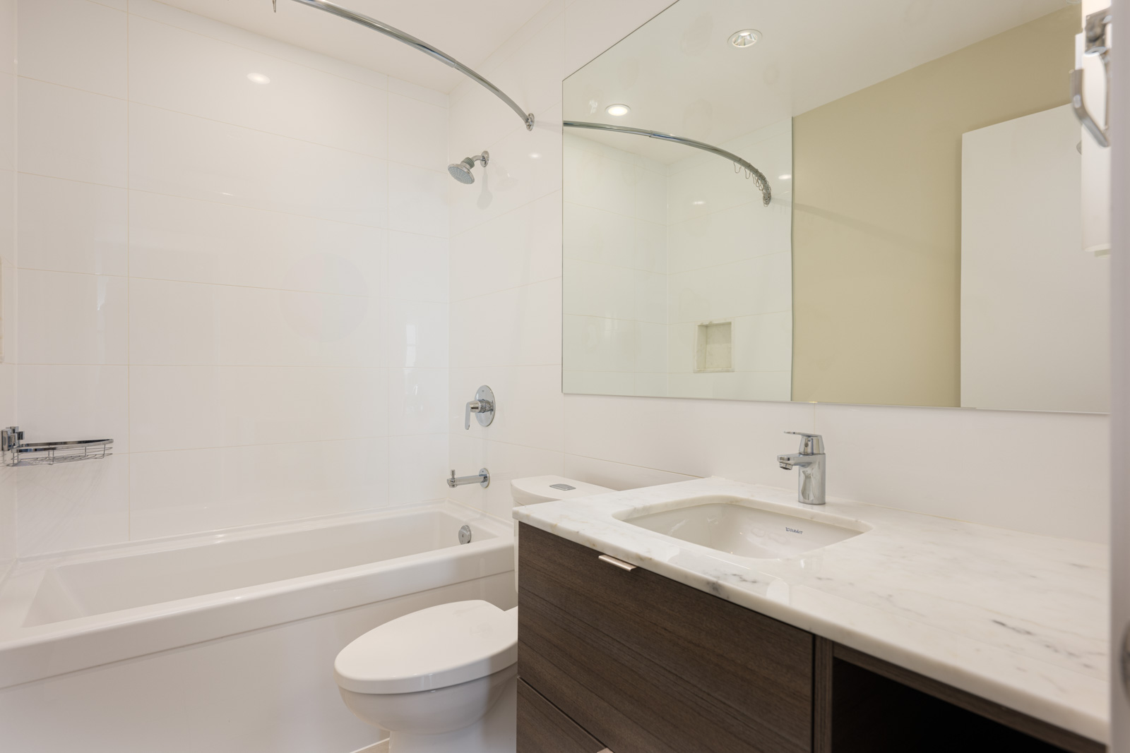 Modern bathroom with a bathtub-shower combo, white tiles, a large mirror, and a dark wood vanity with a white marble countertop and sink.