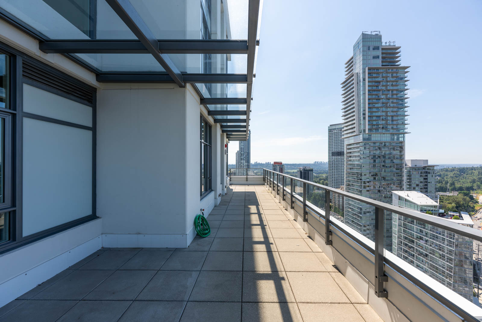 High-rise balcony with glass railing and tiled floor, overlooking modern city buildings under a clear blue sky. A green hose is coiled near the wall.