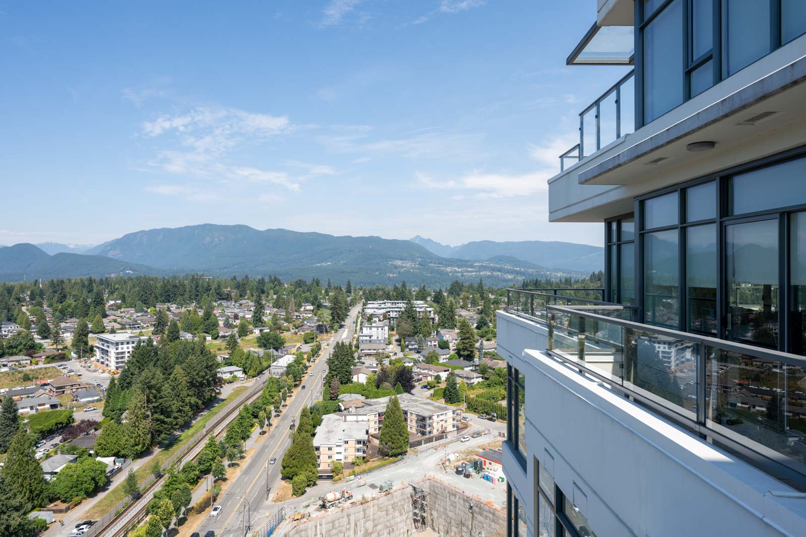 View from a high-rise balcony overlooking a suburban area with roads, houses, trees, and mountains in the distance under a clear sky.