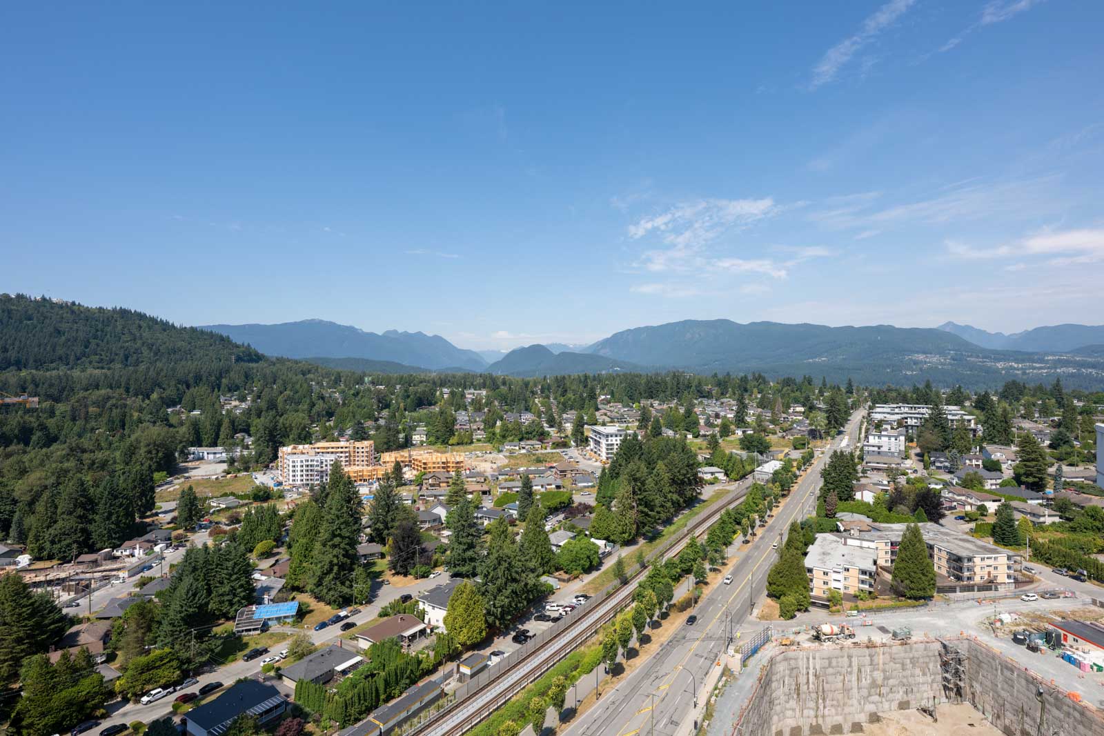 Aerial view of a suburban neighborhood with tree-lined streets, residential buildings, a construction site, and mountains in the background under a clear sky.