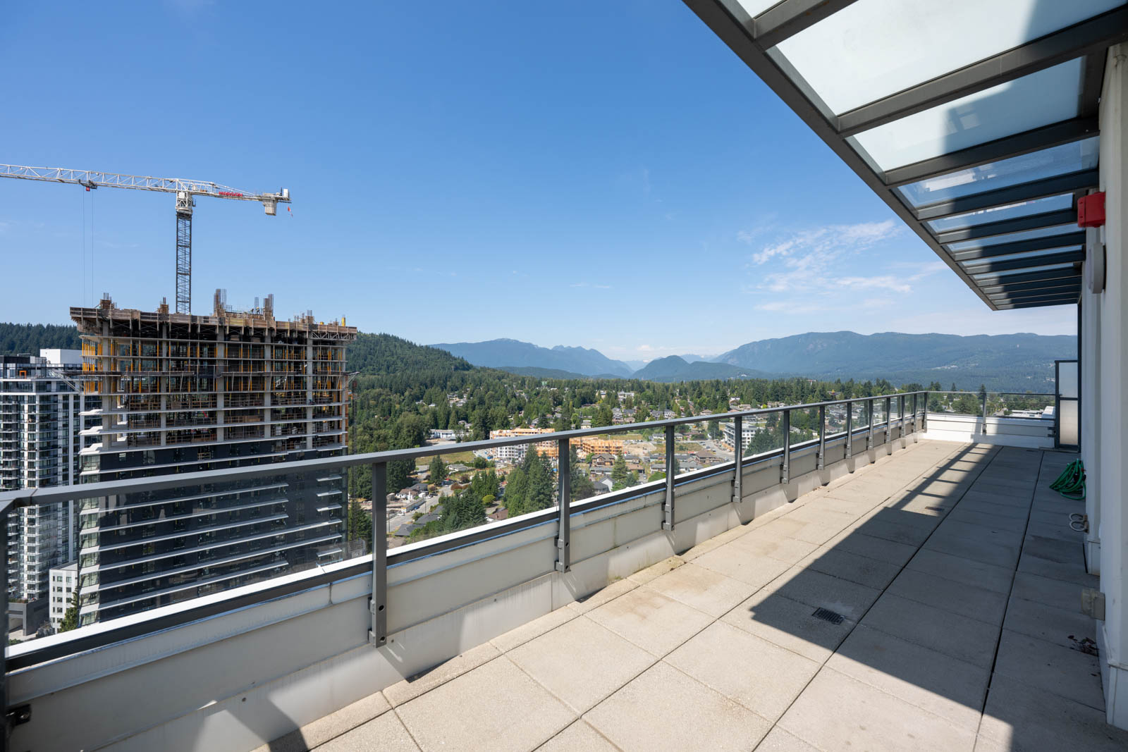 Rooftop terrace with glass railing overlooks a cityscape, construction site, green hills, and distant mountains under a clear blue sky.