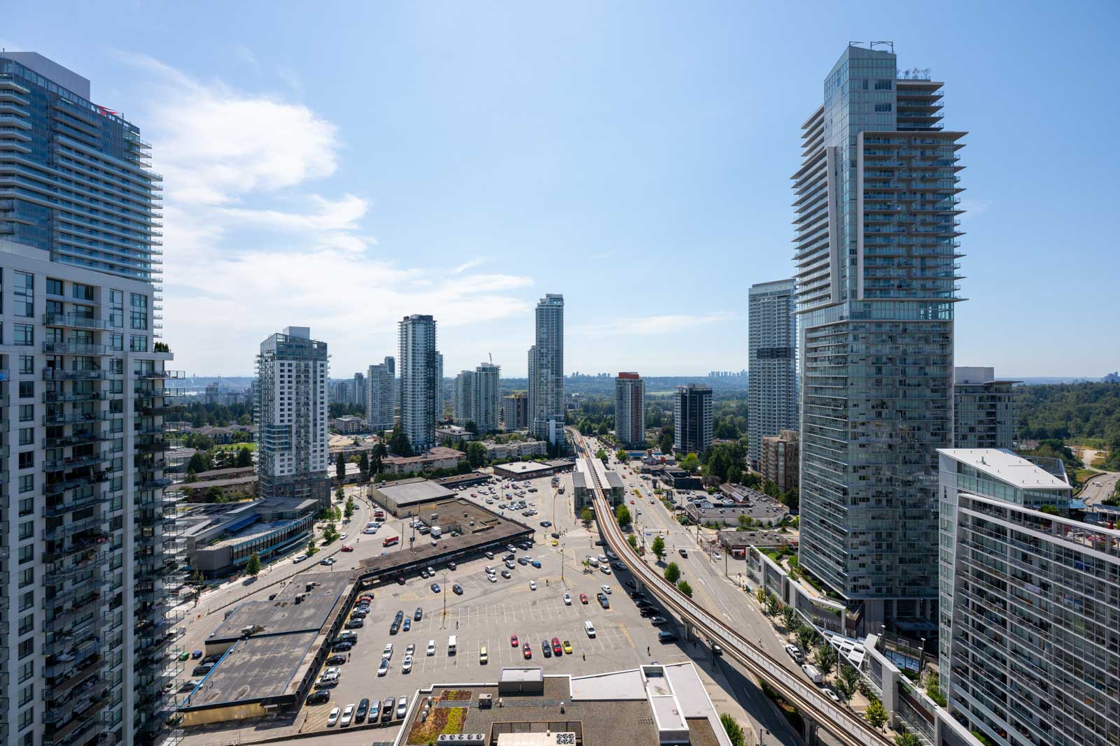A cityscape view of high-rise buildings, a parking lot with cars, and a major road running through the center under a clear sky.