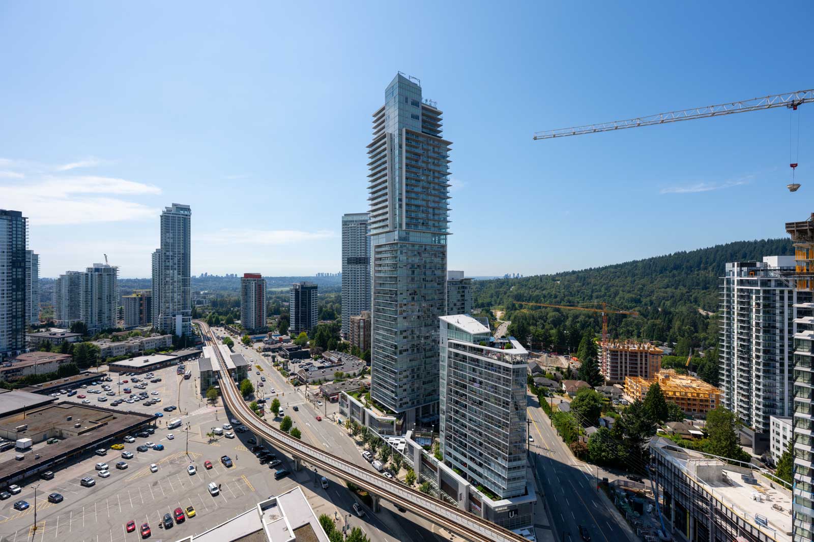 A cityscape featuring tall modern buildings, a construction crane, busy roads, and a forested hillside under a clear blue sky.