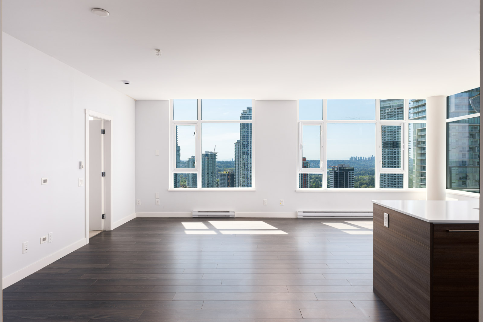Unfurnished modern apartment interior with large windows, dark wood flooring, white walls, and a kitchen island, overlooking a cityscape.