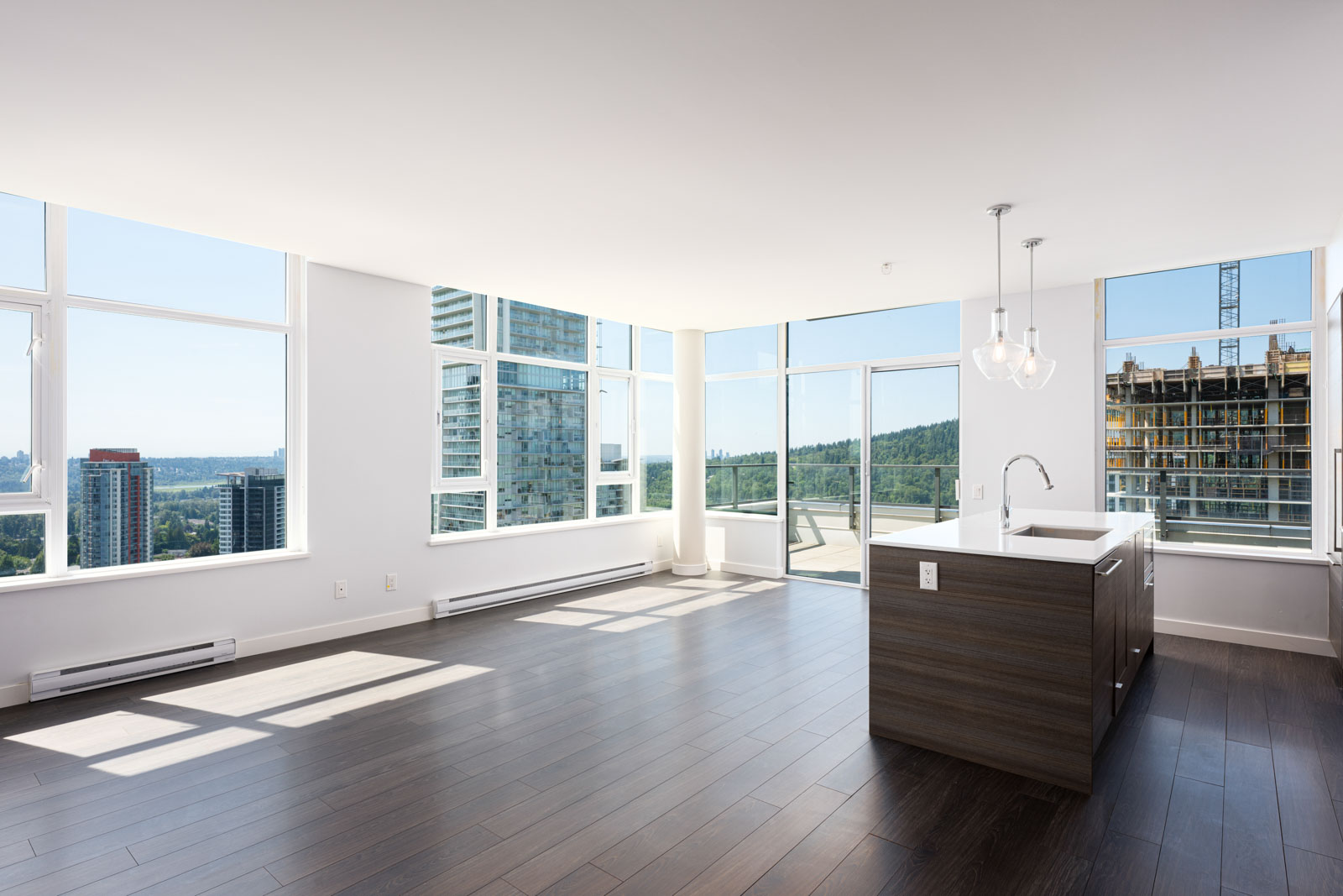 Modern, unfurnished high-rise apartment with large windows, dark wood floors, a kitchen island, and city views visible outside.