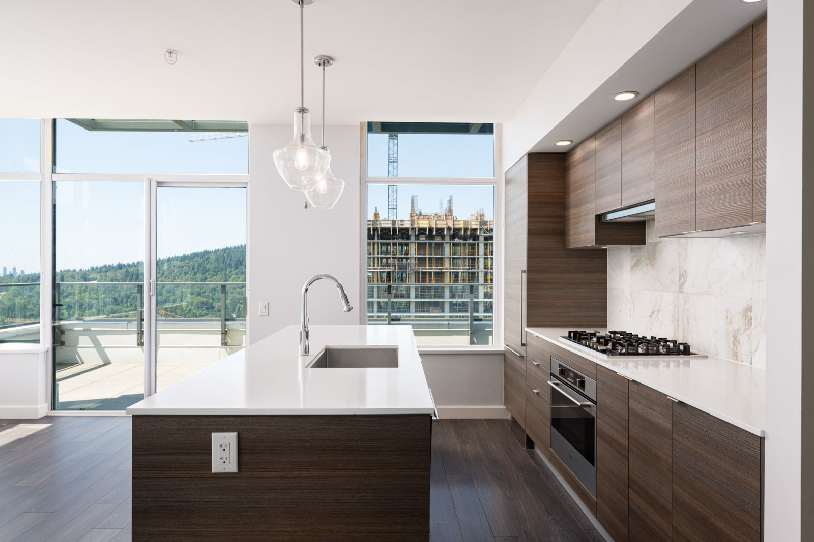 Modern kitchen with wood cabinets, white countertops, and stainless steel appliances, overlooking a balcony with views of trees and a nearby building under construction.
