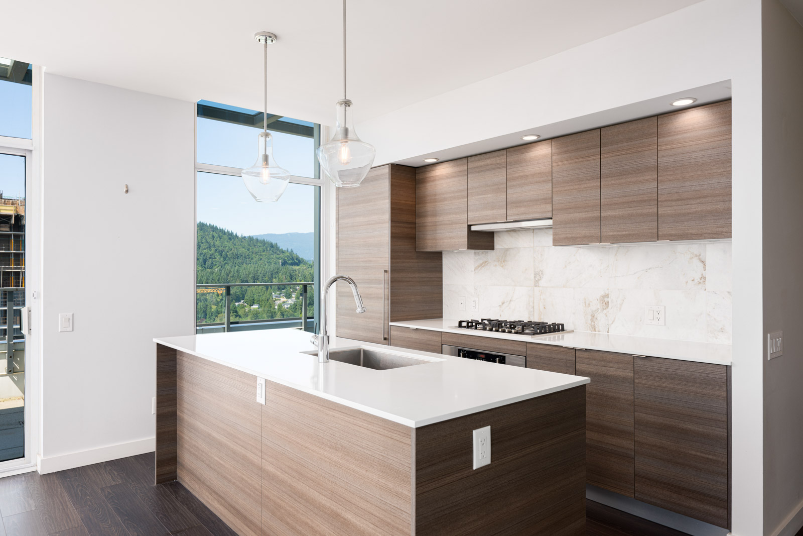 Modern kitchen with wood cabinets, white countertops, a sink island, pendant lights, and large windows showing a view of trees and hills.