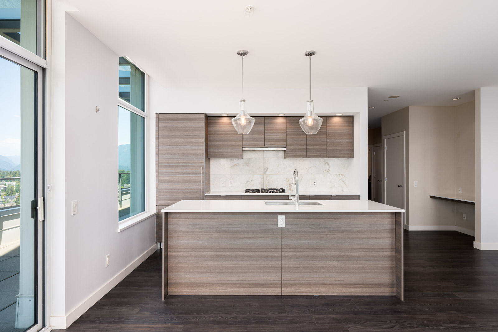 Modern kitchen with wood cabinetry, white countertops, pendant lights, built-in stove, and large windows providing natural light.
