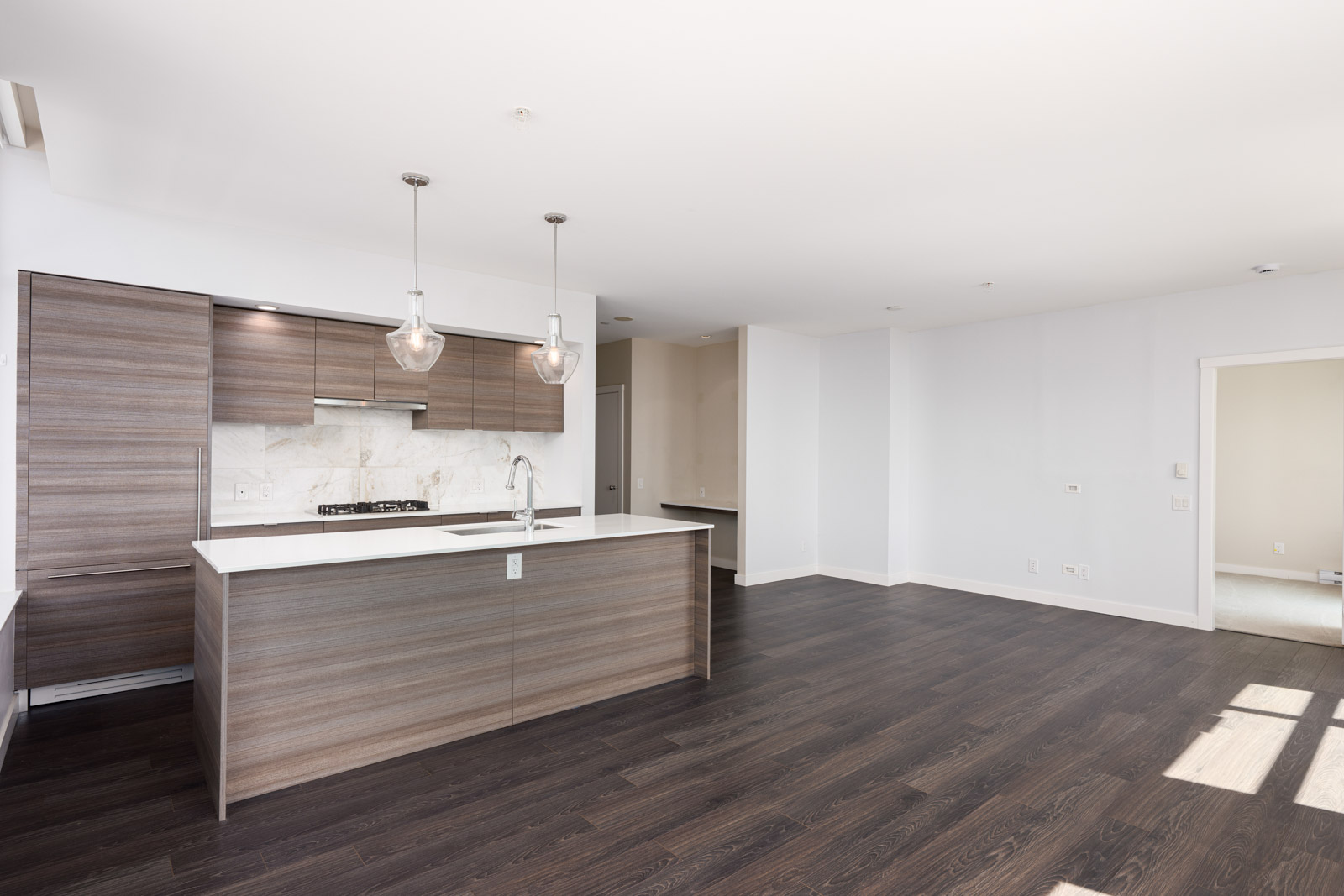 Modern kitchen with wood cabinetry, white countertops, pendant lights, and dark hardwood floors, open to a spacious, sunlit living area with white walls.