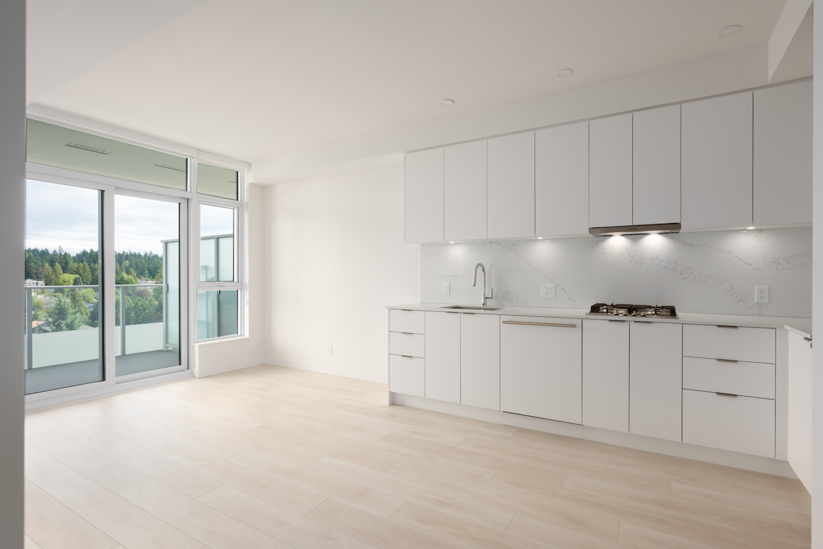 Modern, unfurnished kitchen with white cabinets, built-in stovetop, and dishwasher, adjacent to a bright living area with large windows and balcony overlooking greenery.