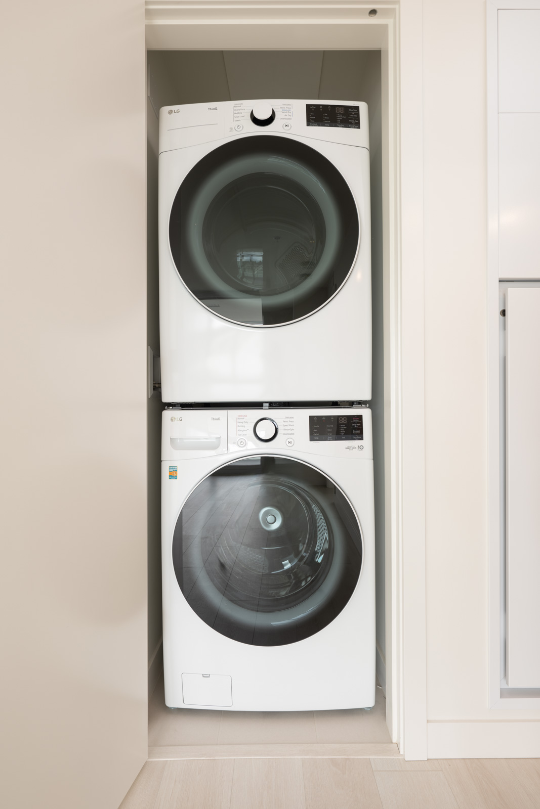 A stacked washer and dryer set in a small, recessed laundry space with white walls and light wood flooring.