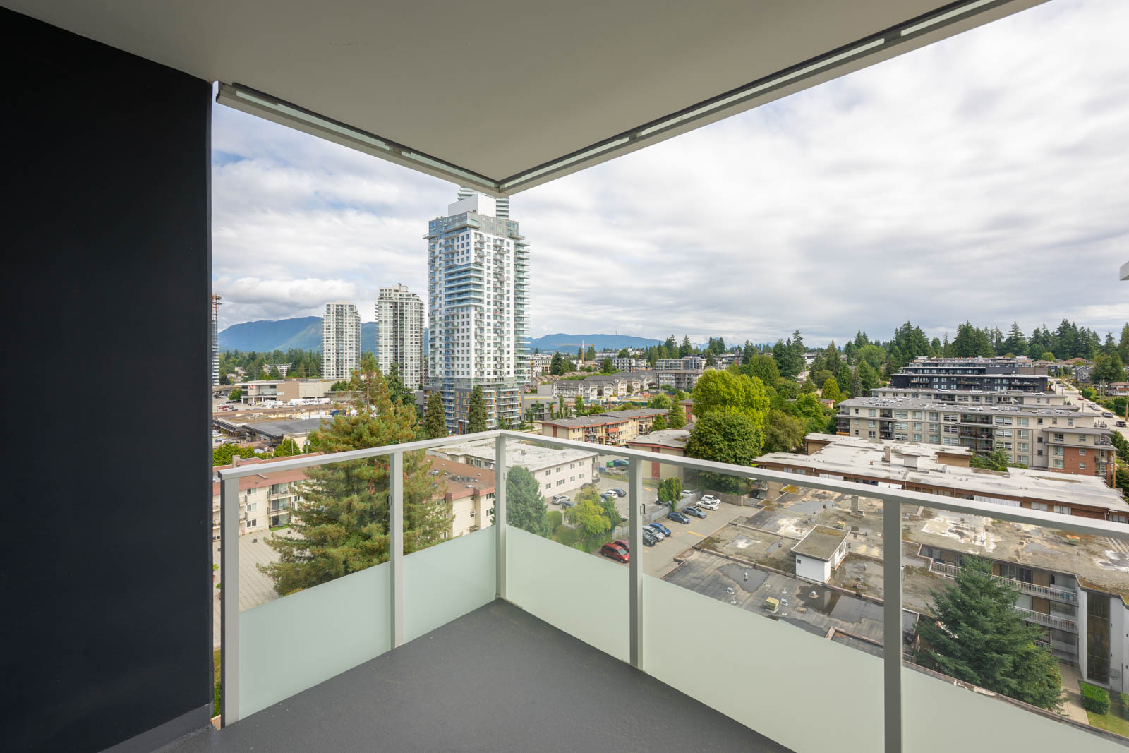 A cityscape view from a high-rise balcony with glass railing, overlooking buildings, trees, and distant mountains under a cloudy sky.