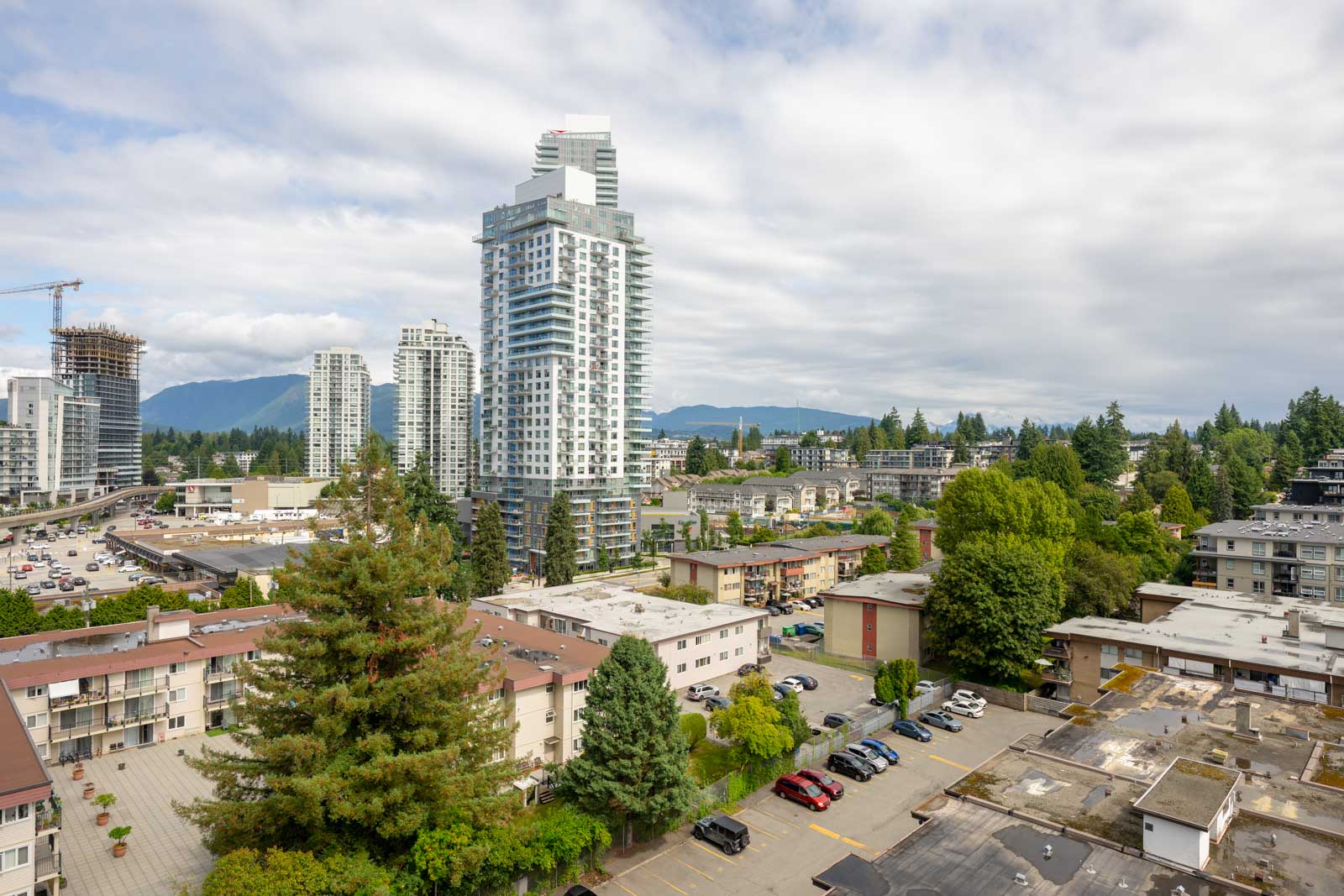A cityscape with residential and high-rise buildings, trees, and distant mountains under a partly cloudy sky.