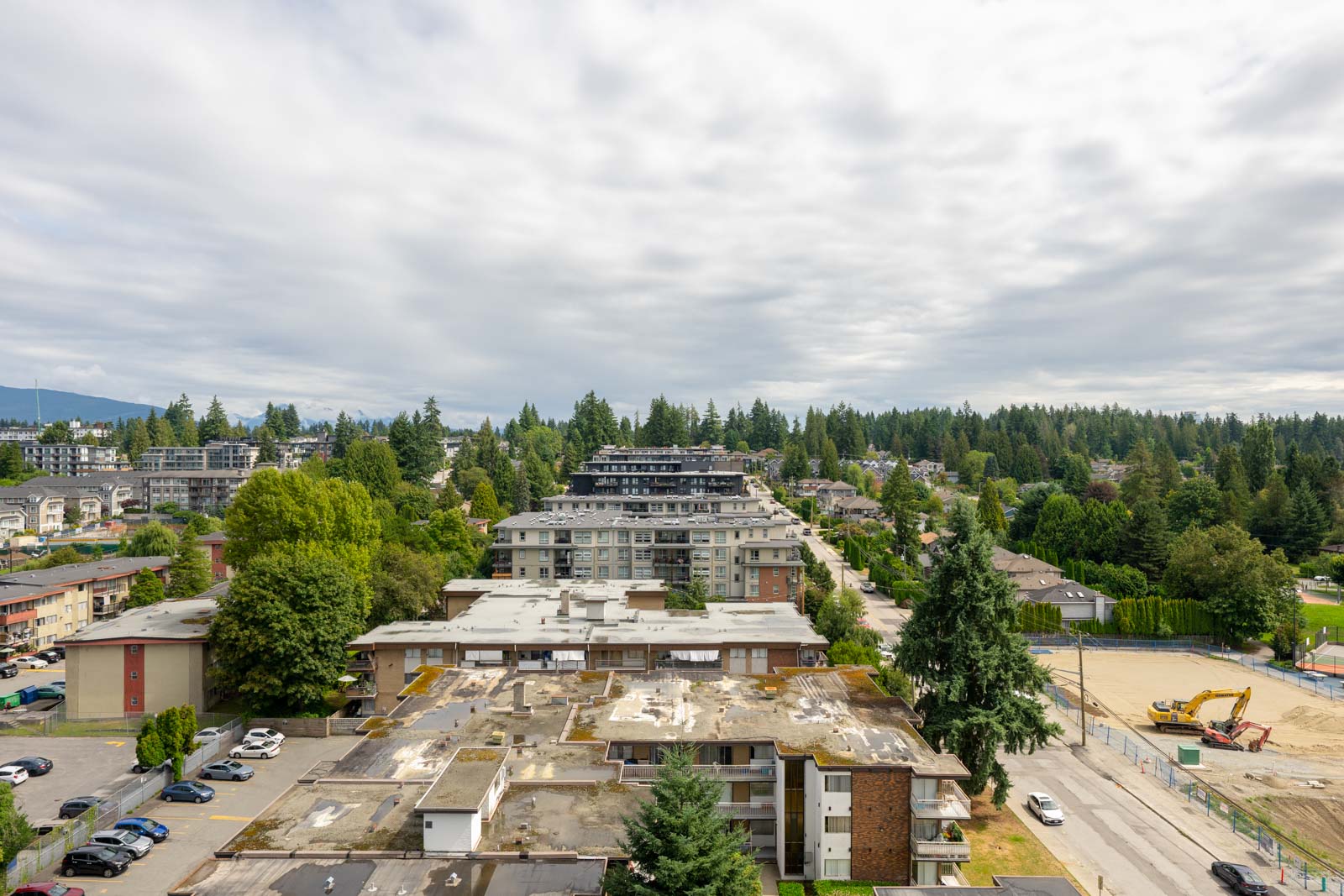 Aerial view of apartment buildings, parking lots, and a construction site surrounded by trees under a cloudy sky.