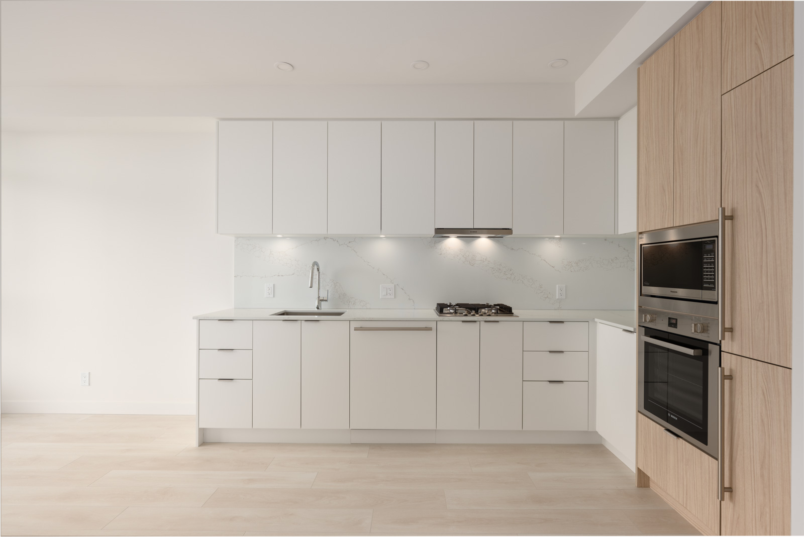 Modern kitchen with white cabinets, built-in oven and microwave, gas stove, sink, and light wood accents. The space is clean, minimalistic, and well-lit.