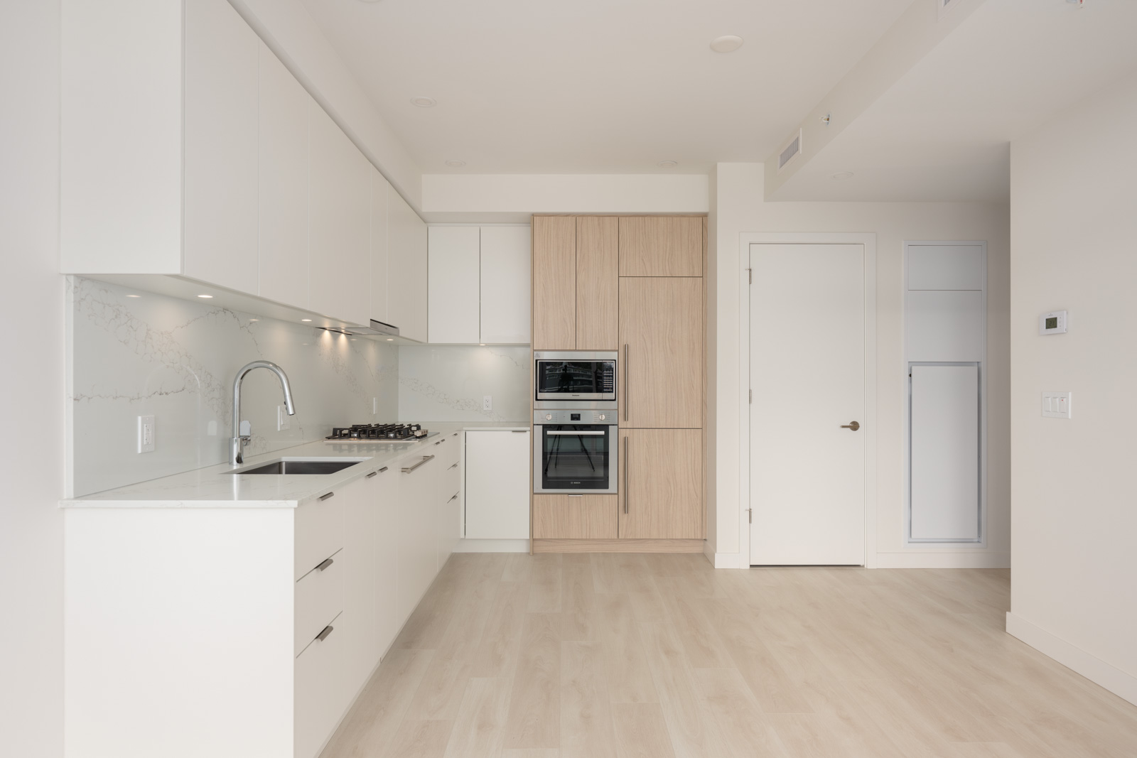 Modern kitchen with white cabinets, light wood finishes, built-in oven, gas cooktop, under-cabinet lighting, and light wood flooring. No furniture or decor present.