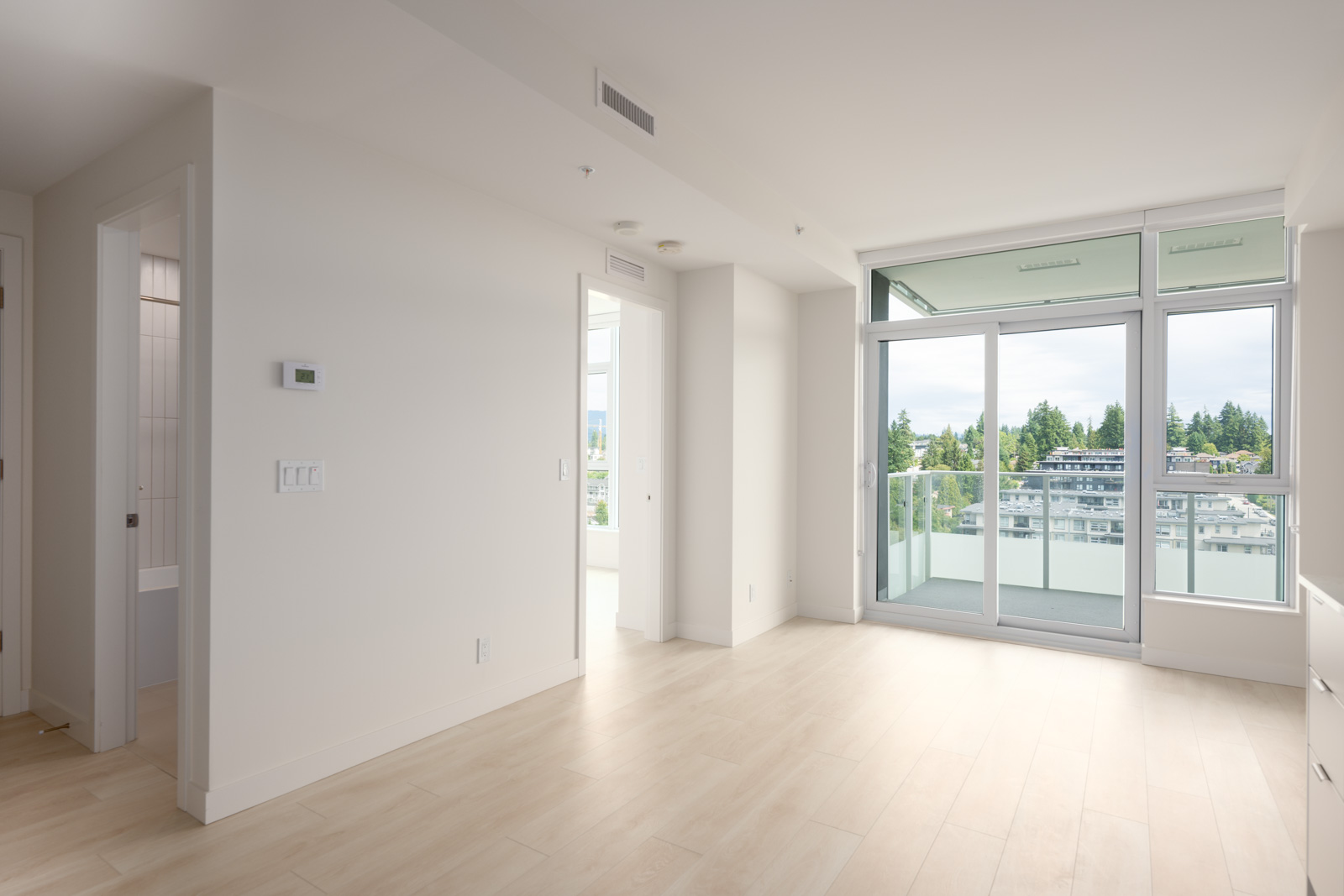 Bright, empty apartment room with light wood flooring, white walls, and large glass doors leading to a balcony with a view of buildings and trees.
