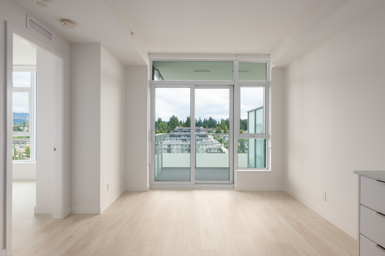Empty, modern apartment room with light wood flooring, large glass doors leading to a balcony, and city views outside. White walls and ample natural light.