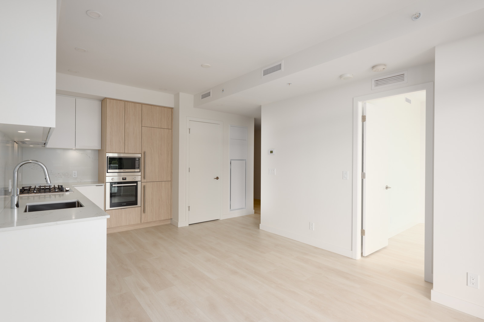 Modern, unfurnished kitchen and living area with light wood floors, white walls, built-in oven, and natural light entering through a doorway to an adjacent room.