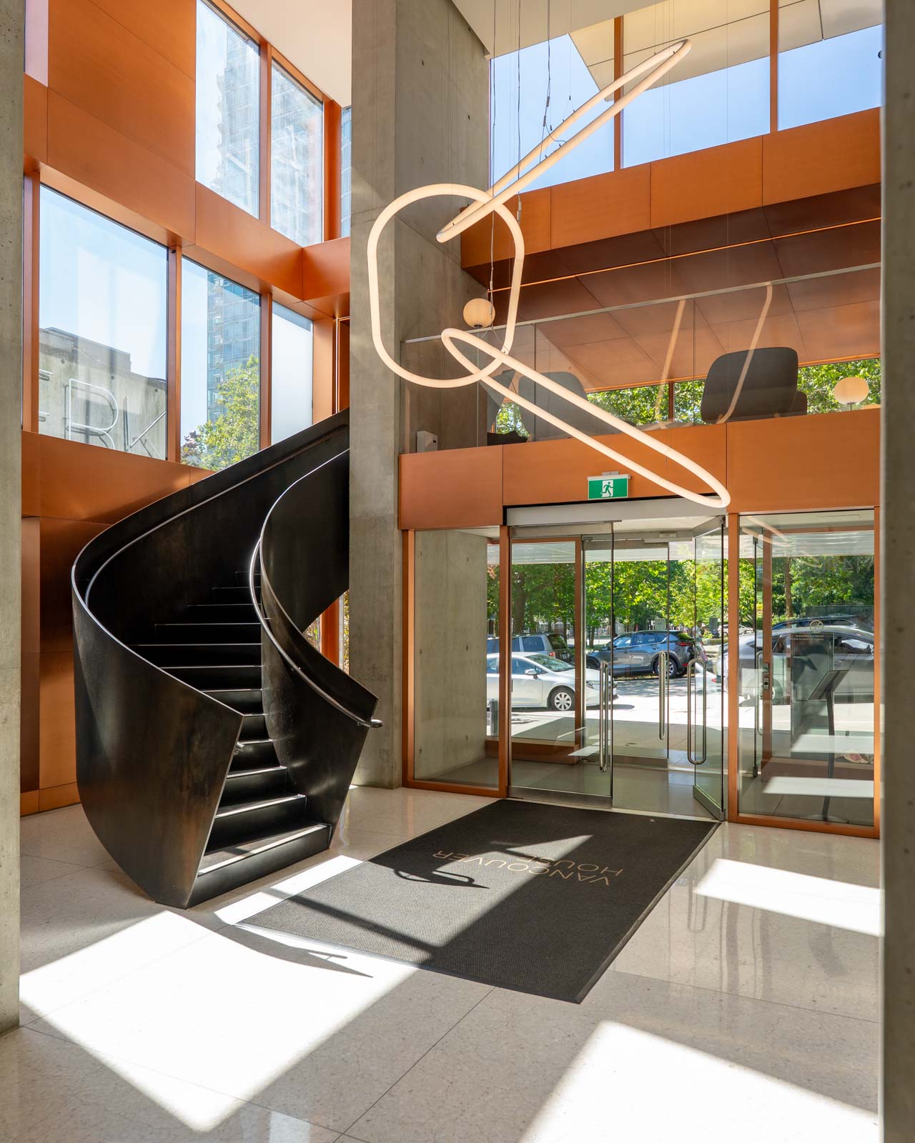 Modern building lobby with a black spiral staircase, large glass windows, geometric light fixture, and a mat labeled 