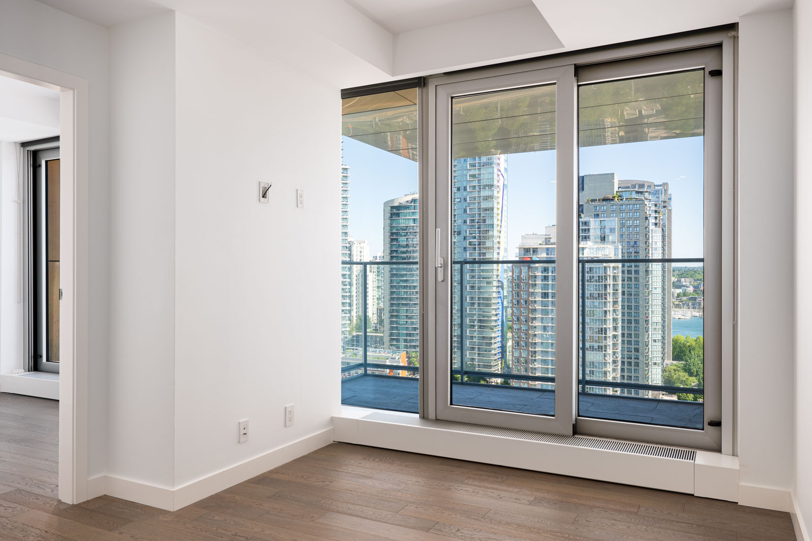 Modern apartment interior with floor-to-ceiling glass doors leading to a balcony, overlooking high-rise buildings and a body of water.