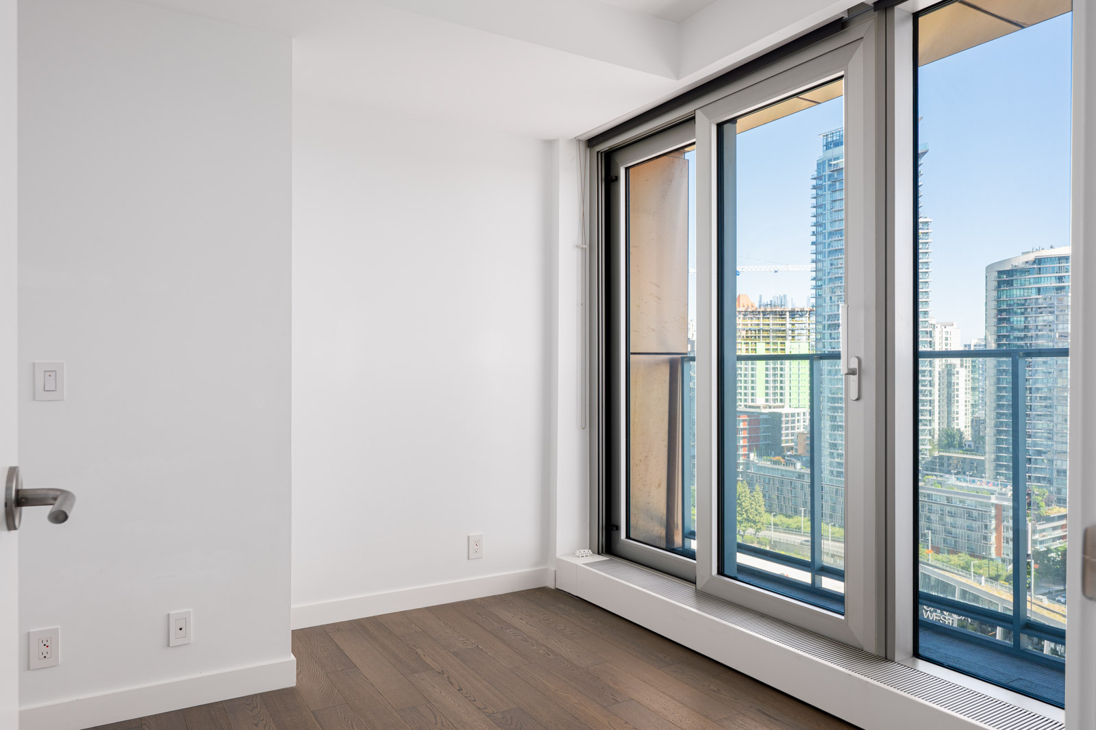 Empty room with white walls, dark wood floor, and a glass door leading to a balcony overlooking modern city buildings.