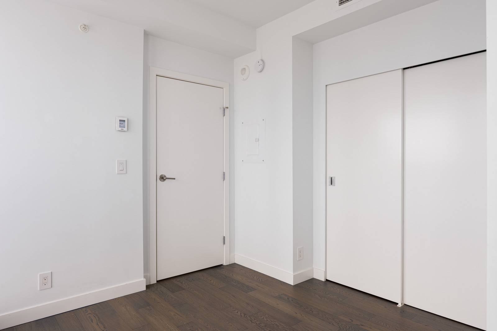 Minimalist room with white walls, a closed white door, a double sliding closet door, a thermostat, and dark wood flooring.