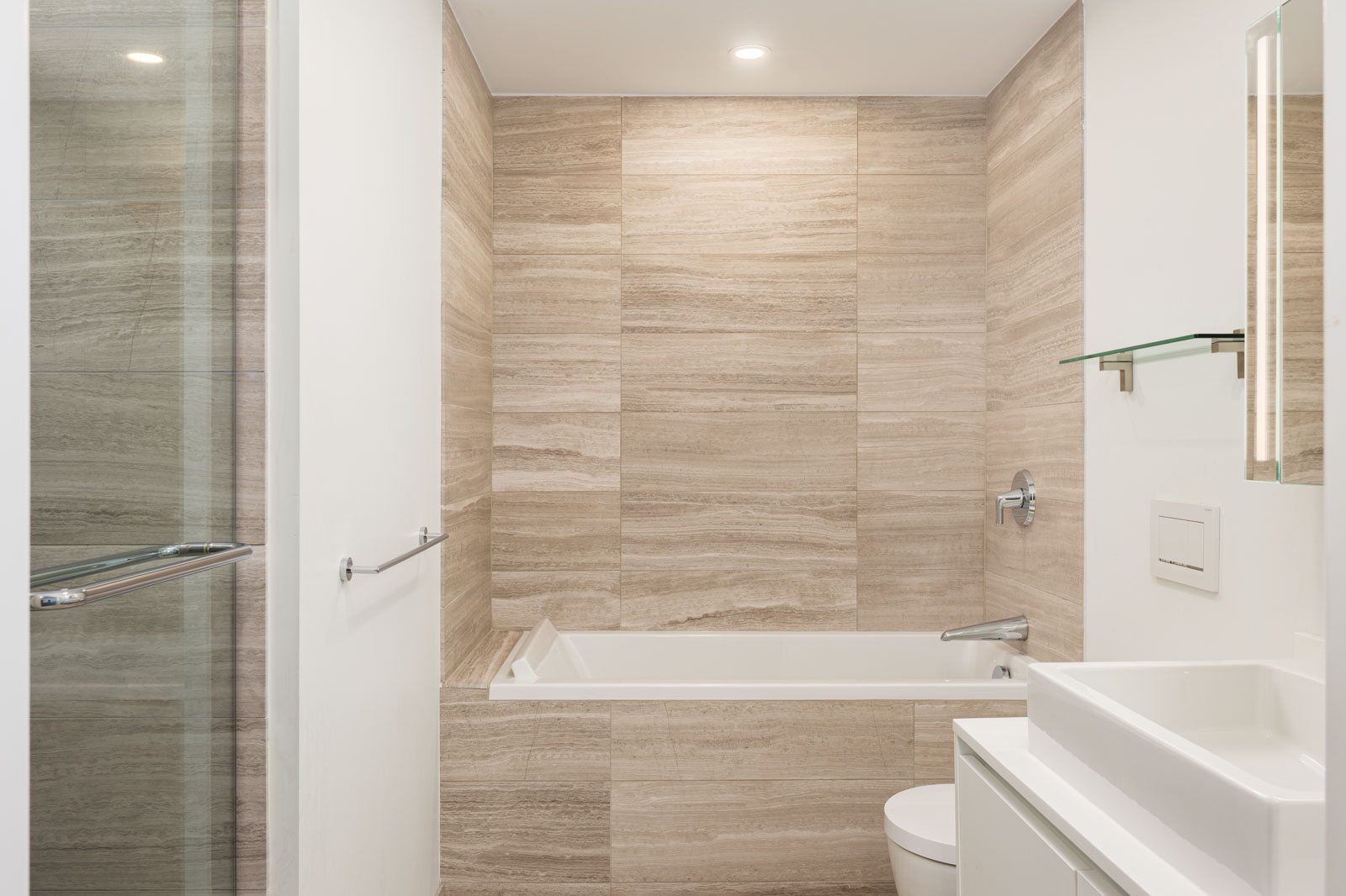 Modern bathroom with beige stone tile walls, a built-in bathtub, a white sink, a toilet, glass shelves, and a glass shower door.