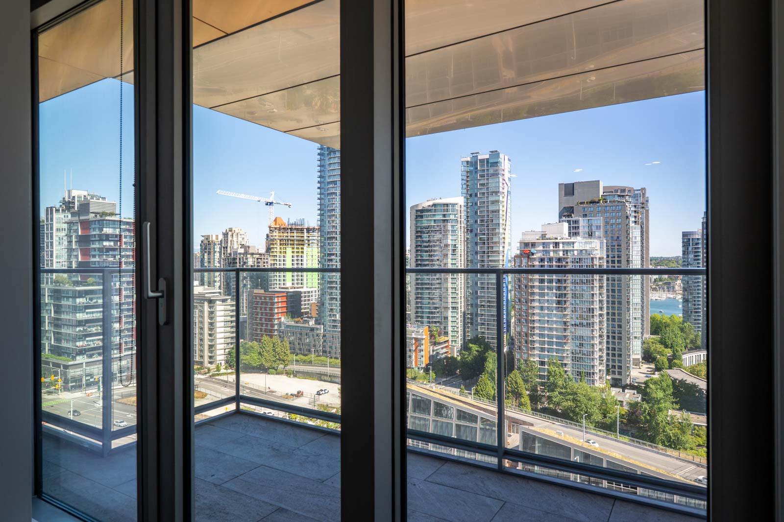 High-rise cityscape view from a balcony with glass railing, seen through floor-to-ceiling windows on a clear day.