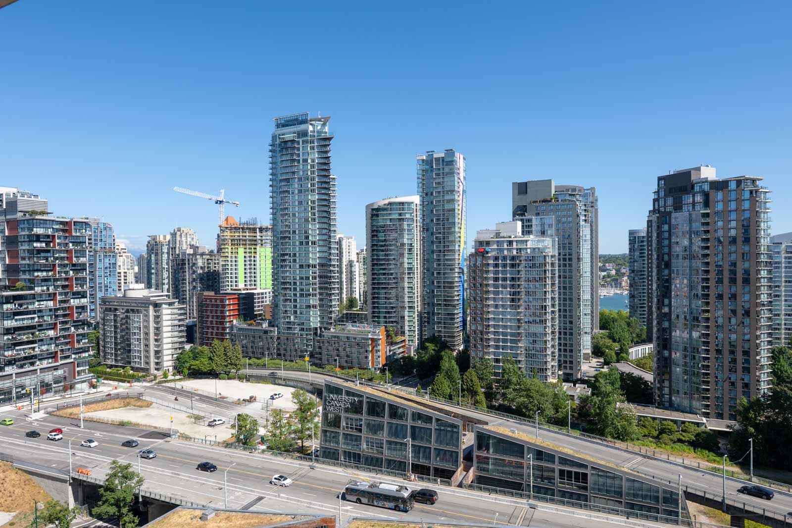 Cityscape with modern high-rise buildings, a few trees, a construction crane, and light traffic on a multi-lane road under a clear blue sky.