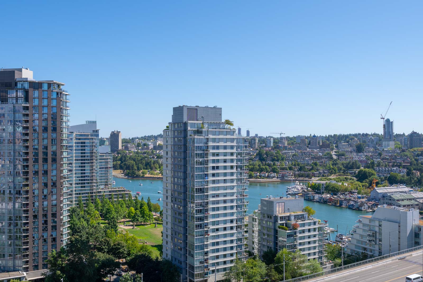 High-rise residential buildings overlook a river with boats, green spaces, and a cityscape in the background under a clear blue sky.