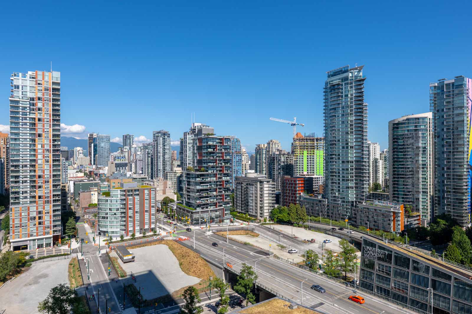 Downtown cityscape with high-rise buildings under a clear blue sky, visible roads, cars, and construction crane in the background.