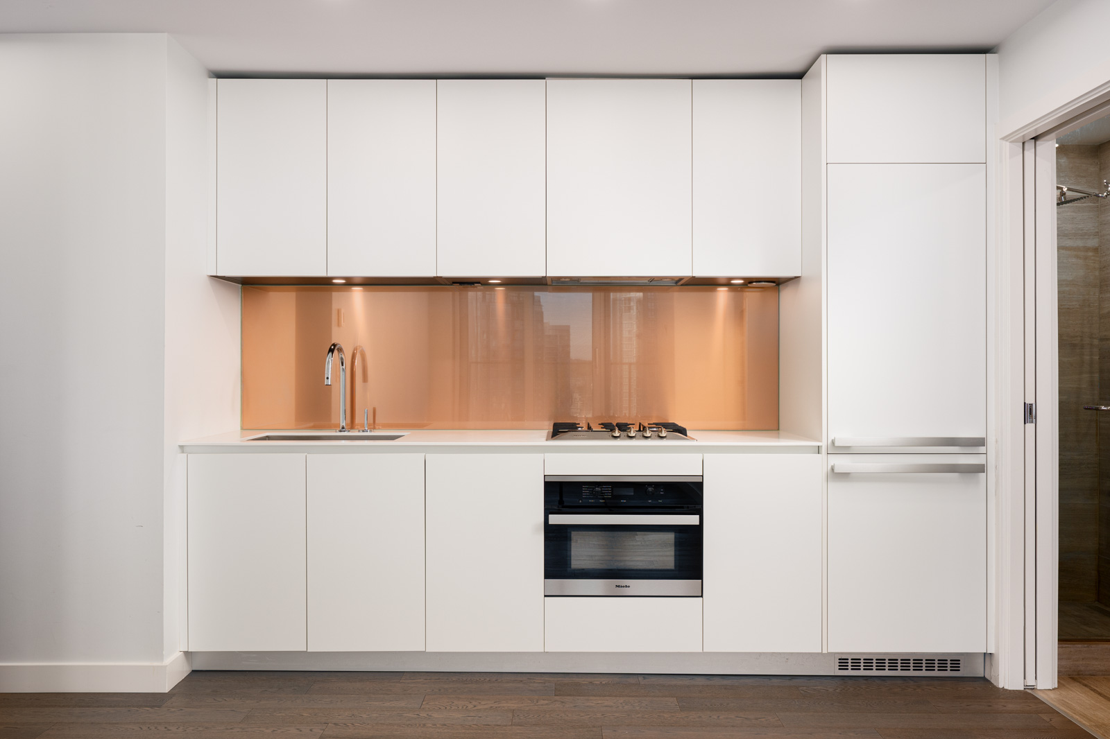 Modern kitchen with white cabinets, a built-in oven, gas cooktop, sink, and a beige backsplash, set against a light wood floor and adjacent to a bathroom doorway.