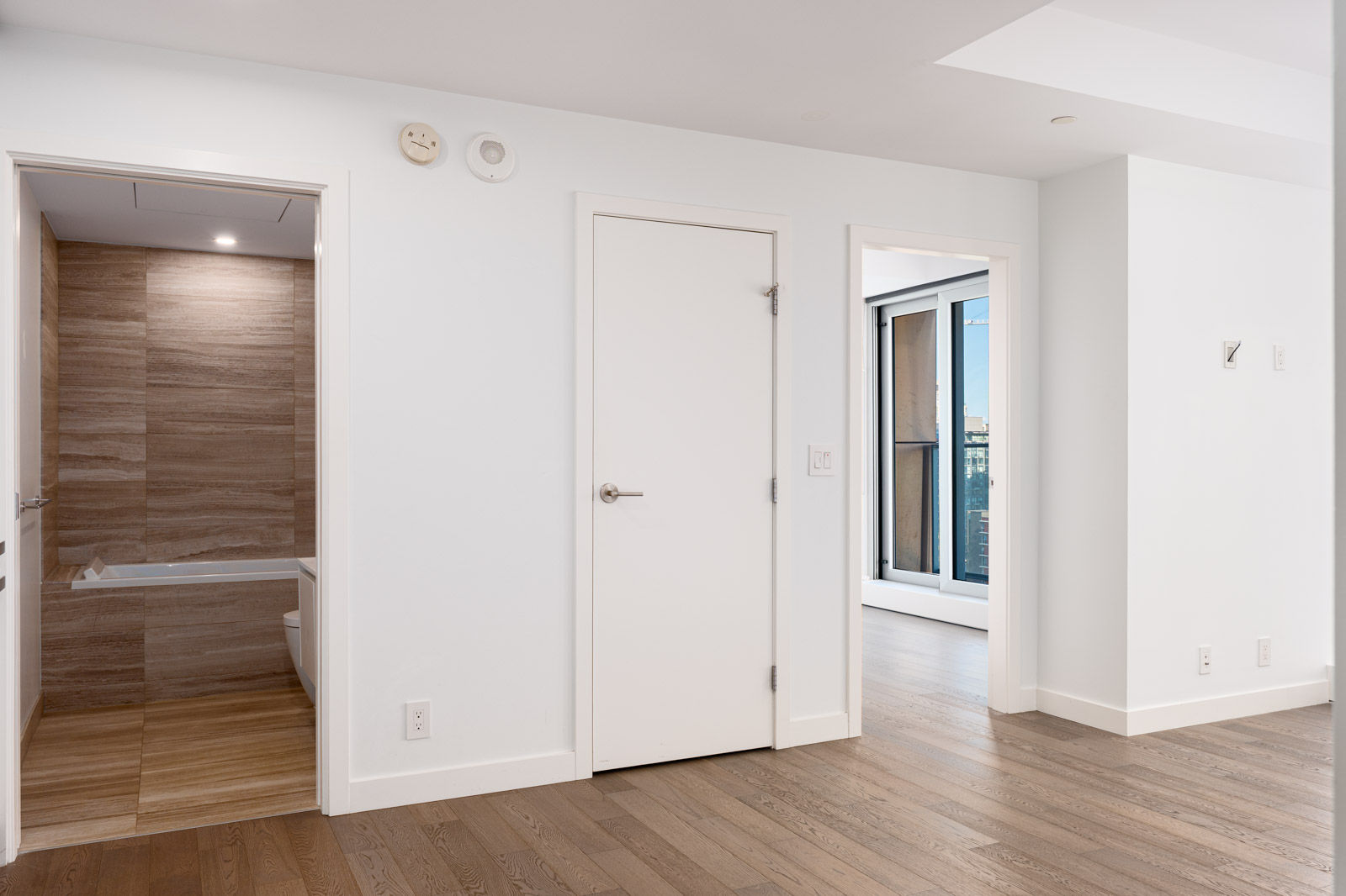 Modern interior with hardwood floors, white walls, a closed door in the center, an open bathroom on the left, and a glass door leading to a balcony on the right.