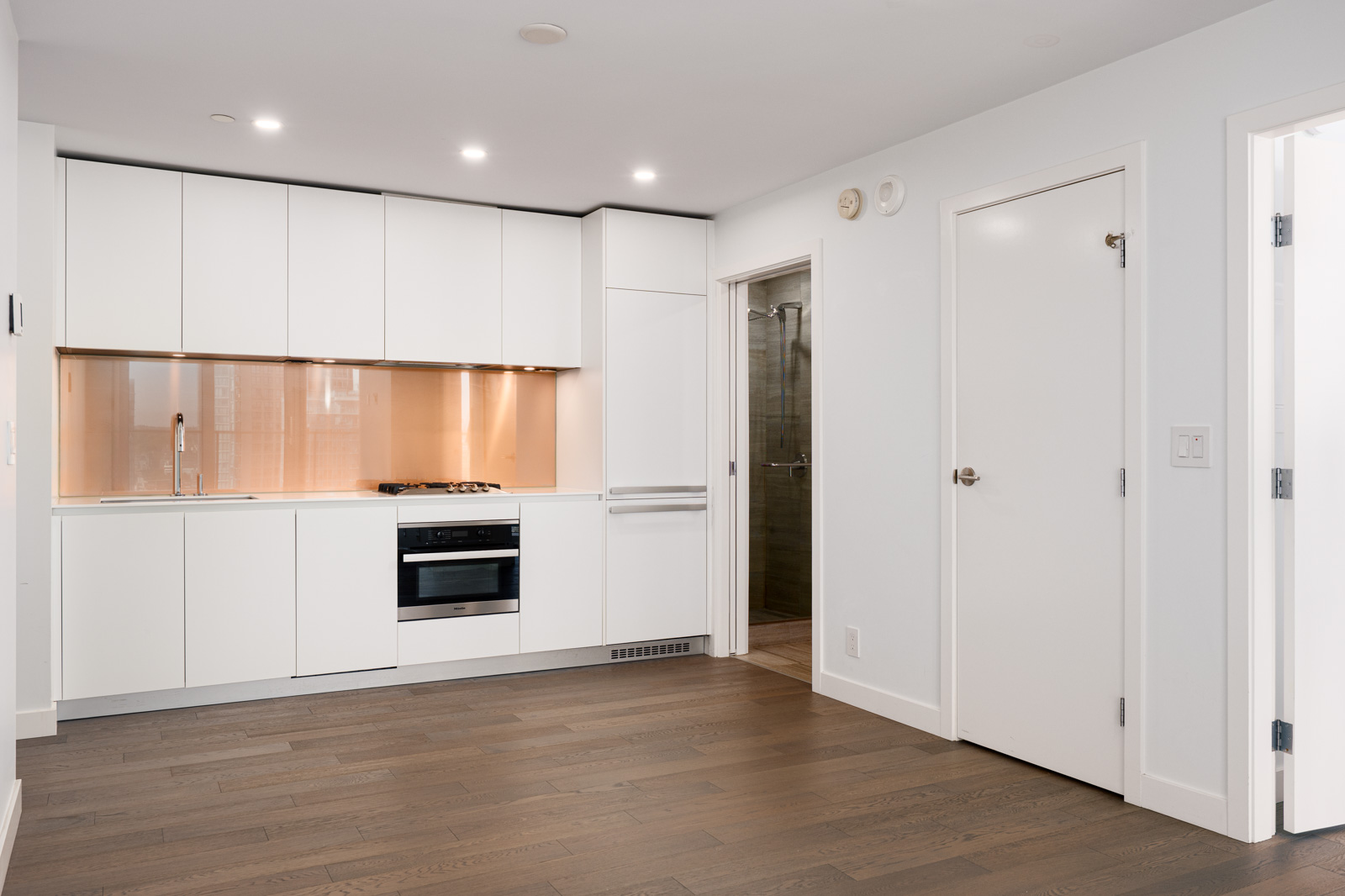 Modern kitchen with white cabinets, built-in oven, and stovetop, featuring a light brown backsplash and dark wood flooring. A door and entry to a bathroom are on the right.