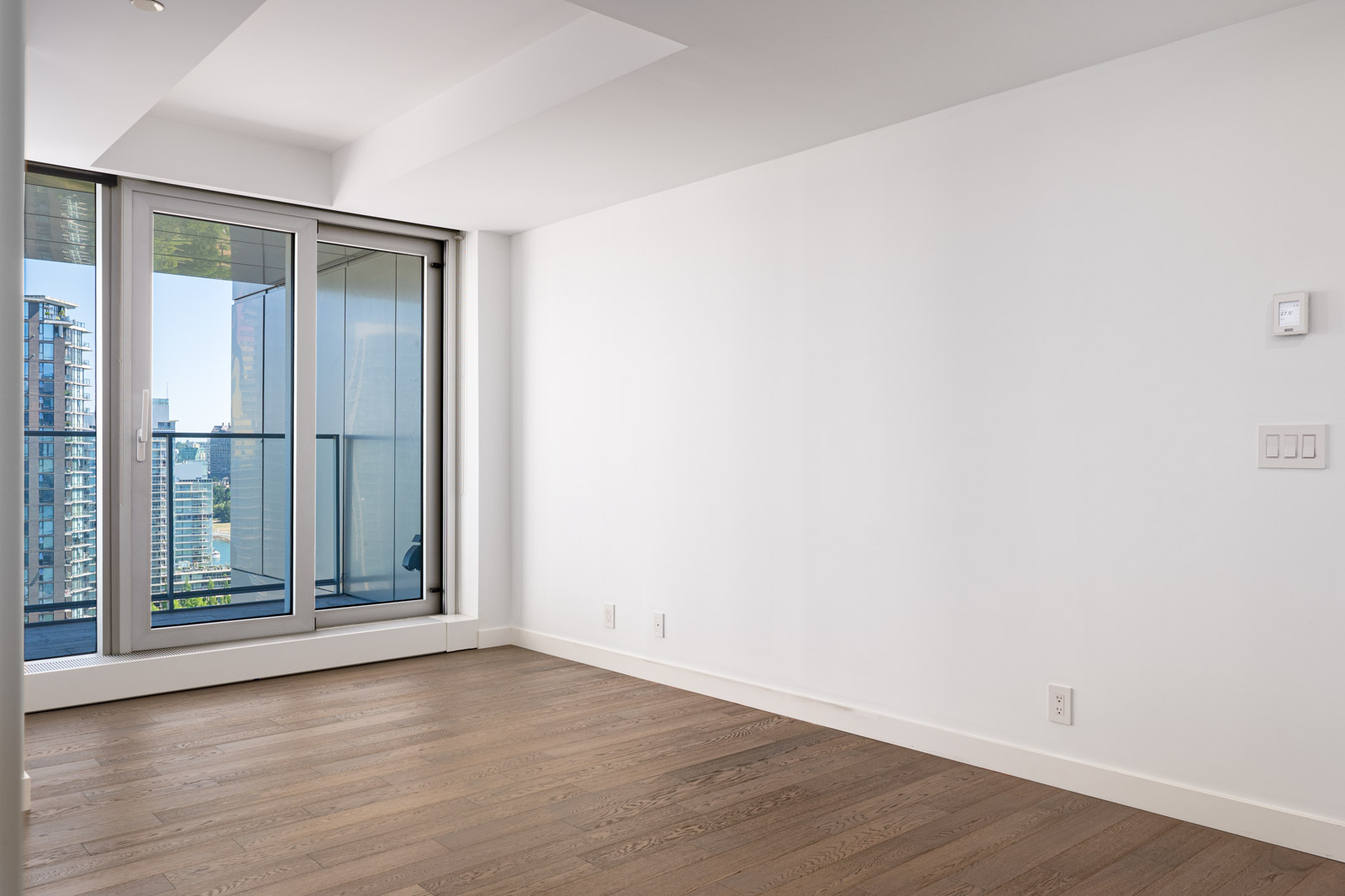 Empty modern room with light wood flooring, white walls, and floor-to-ceiling windows leading to a balcony with a city view.