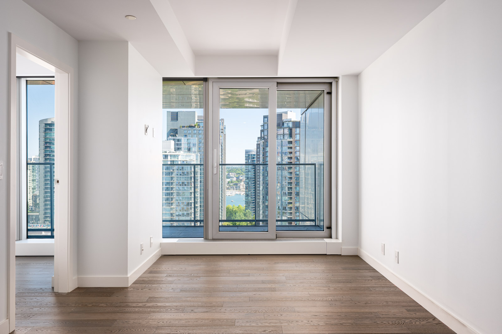 Empty modern apartment room with hardwood floors, white walls, large glass door to balcony, and view of city buildings and water outside.