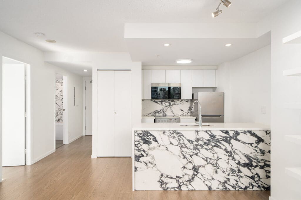 Modern kitchen with white cabinets, stainless steel appliances, and prominent black and white marble surfaces on the island and backsplash. Wood flooring extends into the hallway.