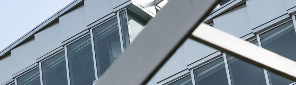 Close-up view of a modern building facade with large windows, partially obscured by intersecting metal beams in the foreground.