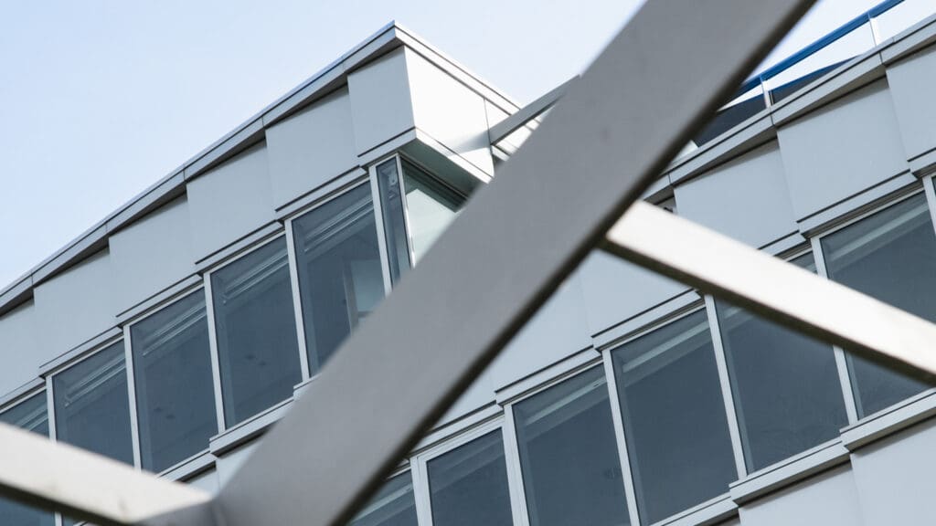 Close-up view of a modern building facade with large windows, partially obscured by intersecting metal beams in the foreground.
