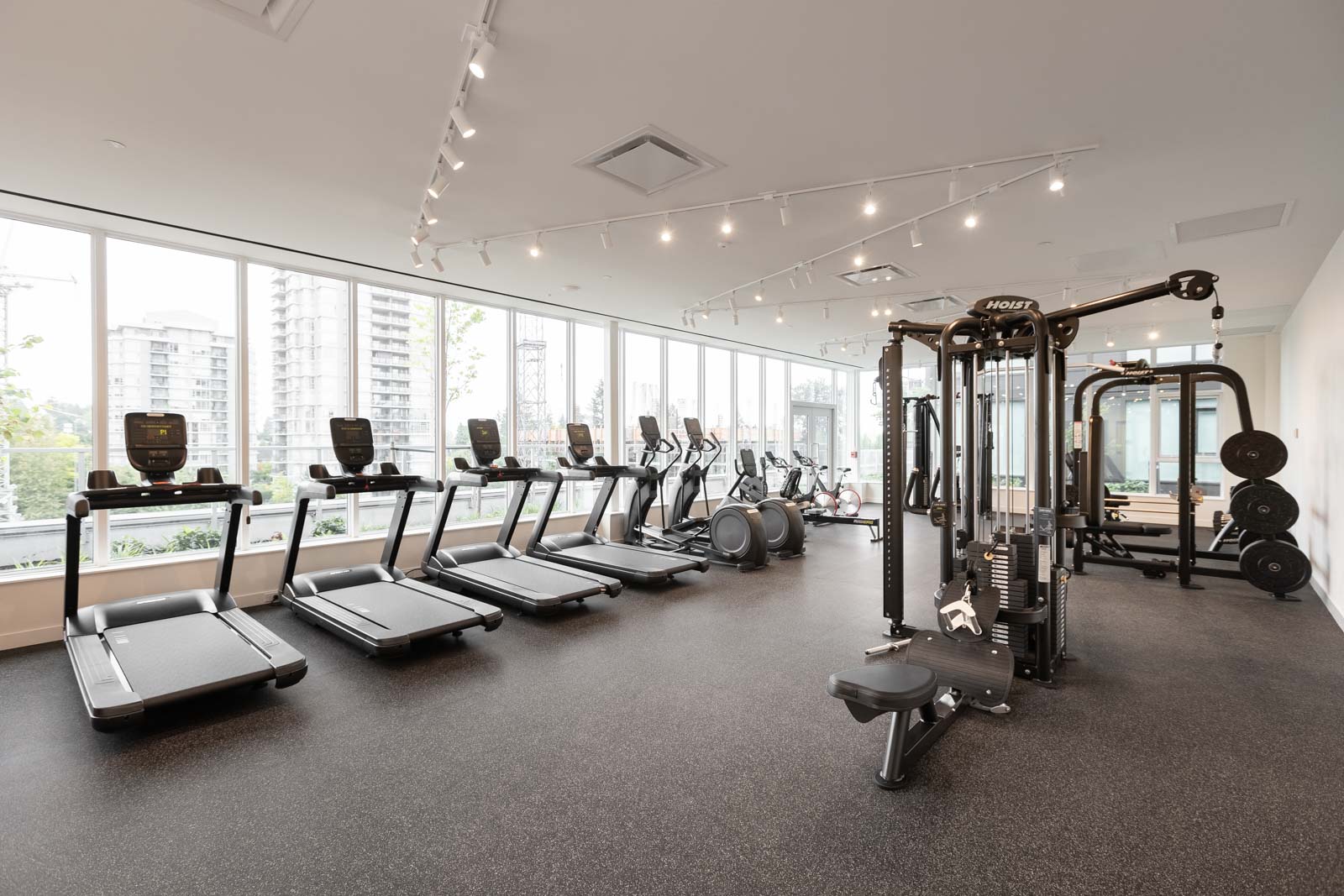 Modern gym with treadmills, stationary bikes, elliptical machines, a multi-purpose fitness station, and weightlifting equipment. Large windows provide natural light and city views.