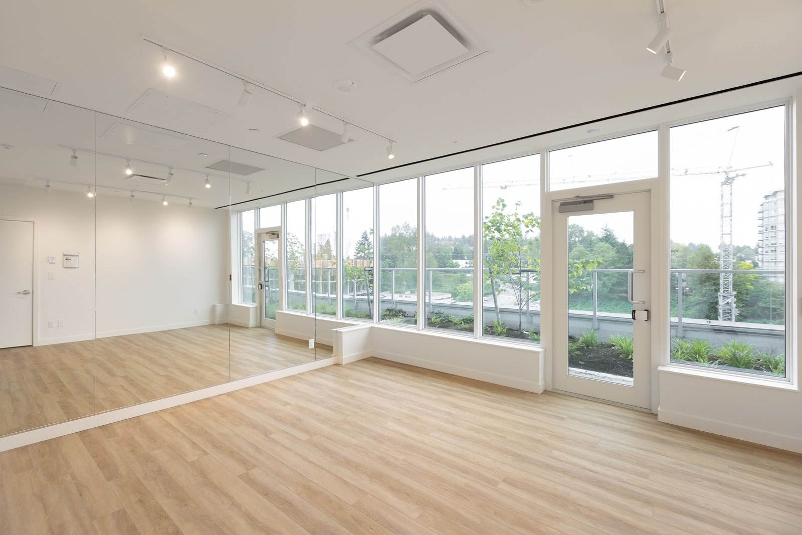 A bright, empty room with wood flooring, floor-to-ceiling windows, a mirrored wall, and a door leading to an outdoor patio with greenery and construction cranes visible outside.