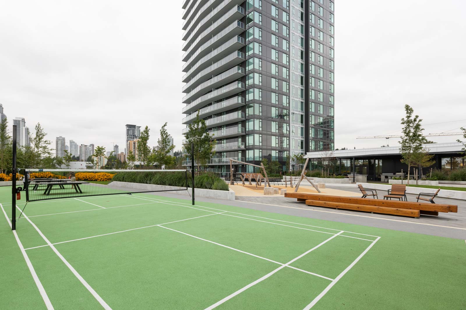 Outdoor green badminton court with net in front of a modern high-rise apartment building, surrounded by landscaped seating and greenery.