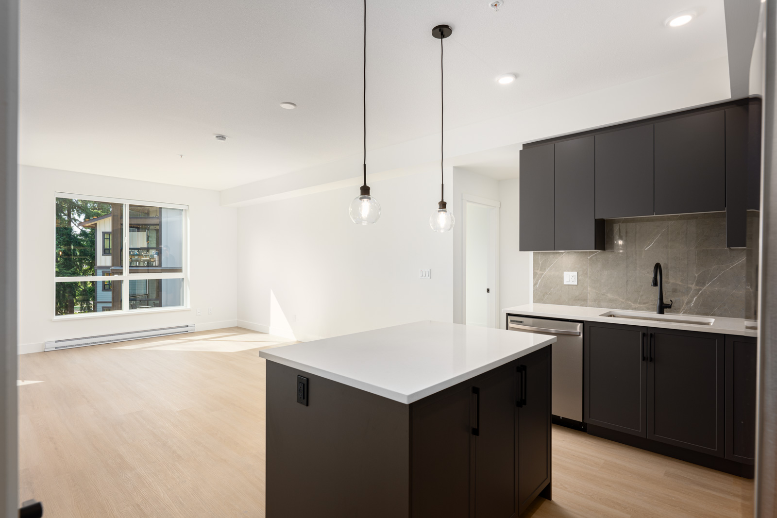 Modern apartment kitchen with dark cabinets, an island with light countertop, pendant lights, and a bright living area with large window in the background.