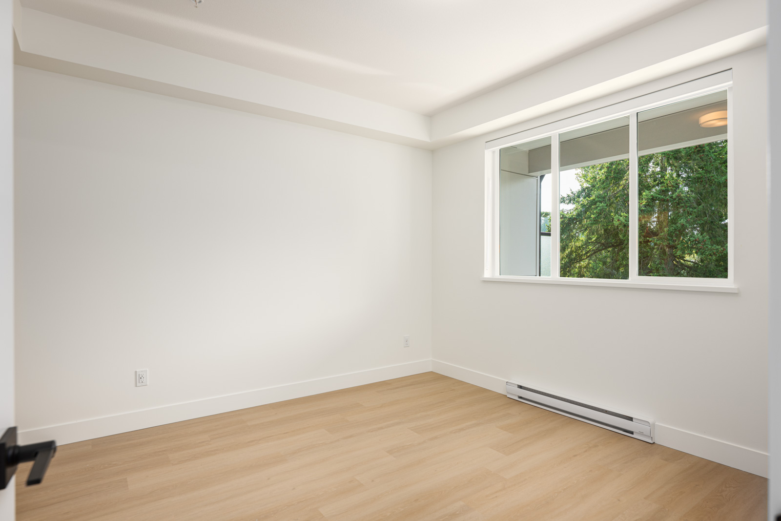 Empty room with light wood flooring, white walls, a large window showing greenery outside, and a baseboard heater below the window.
