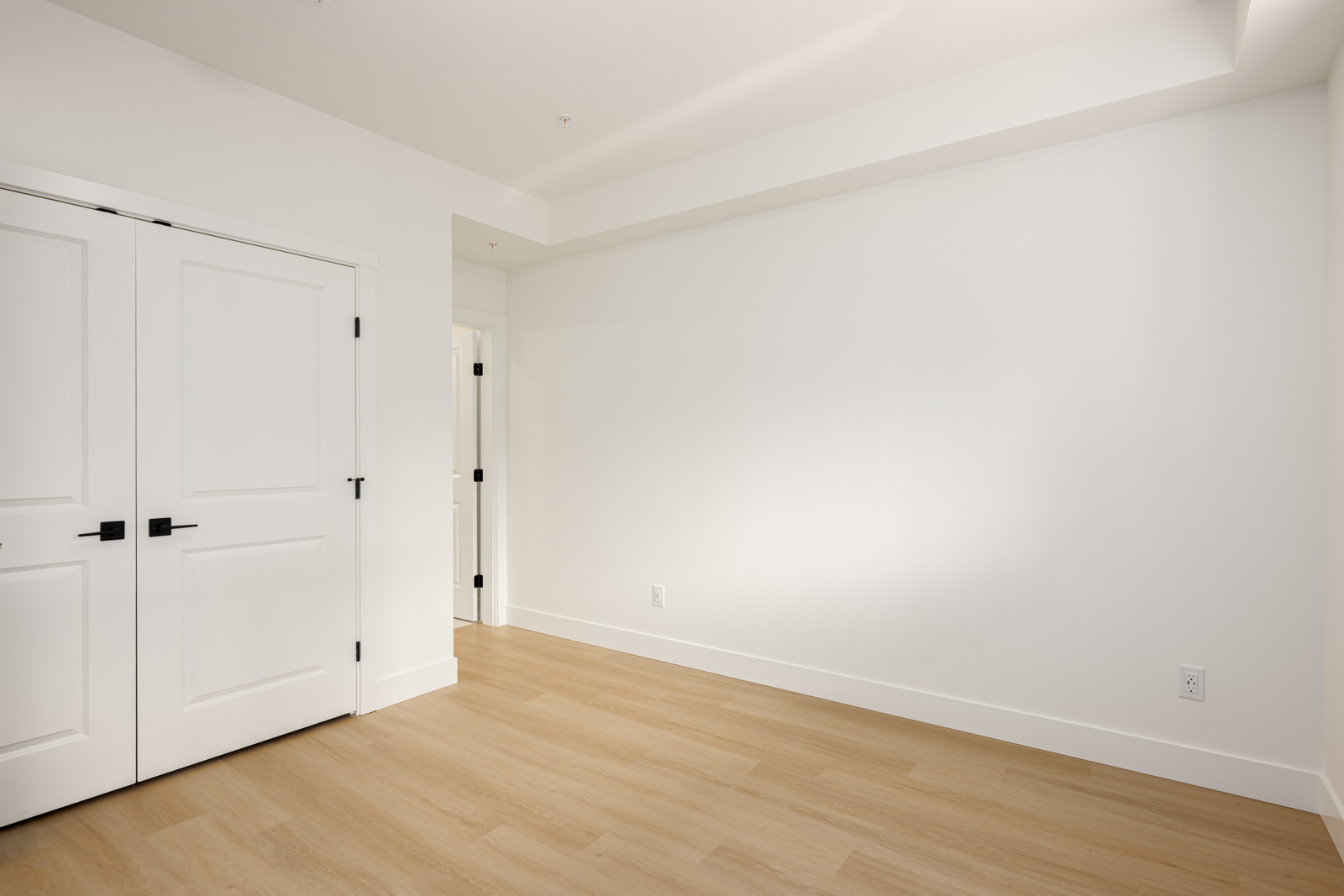 Empty room with light wood flooring, white walls, a double door closet on the left, and a single door leading to another area.