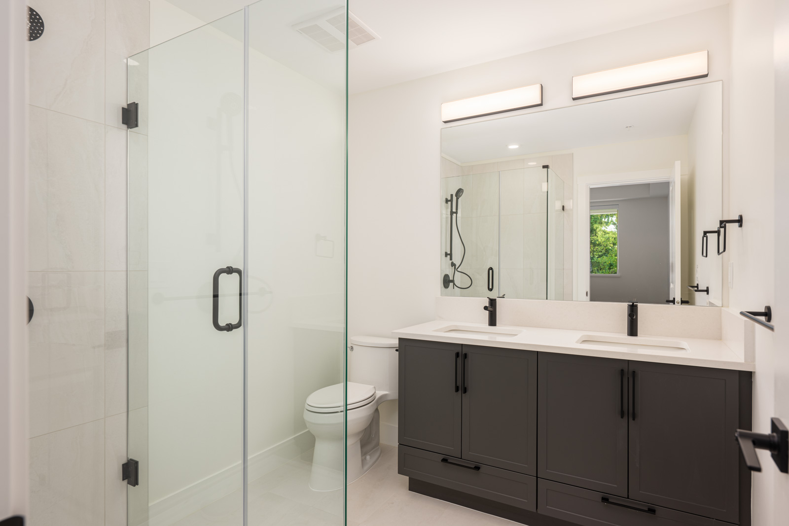 Modern bathroom with a glass shower enclosure, double sink vanity with dark cabinets, two mirrors, and a toilet. The room has bright lighting and a window with greenery visible.