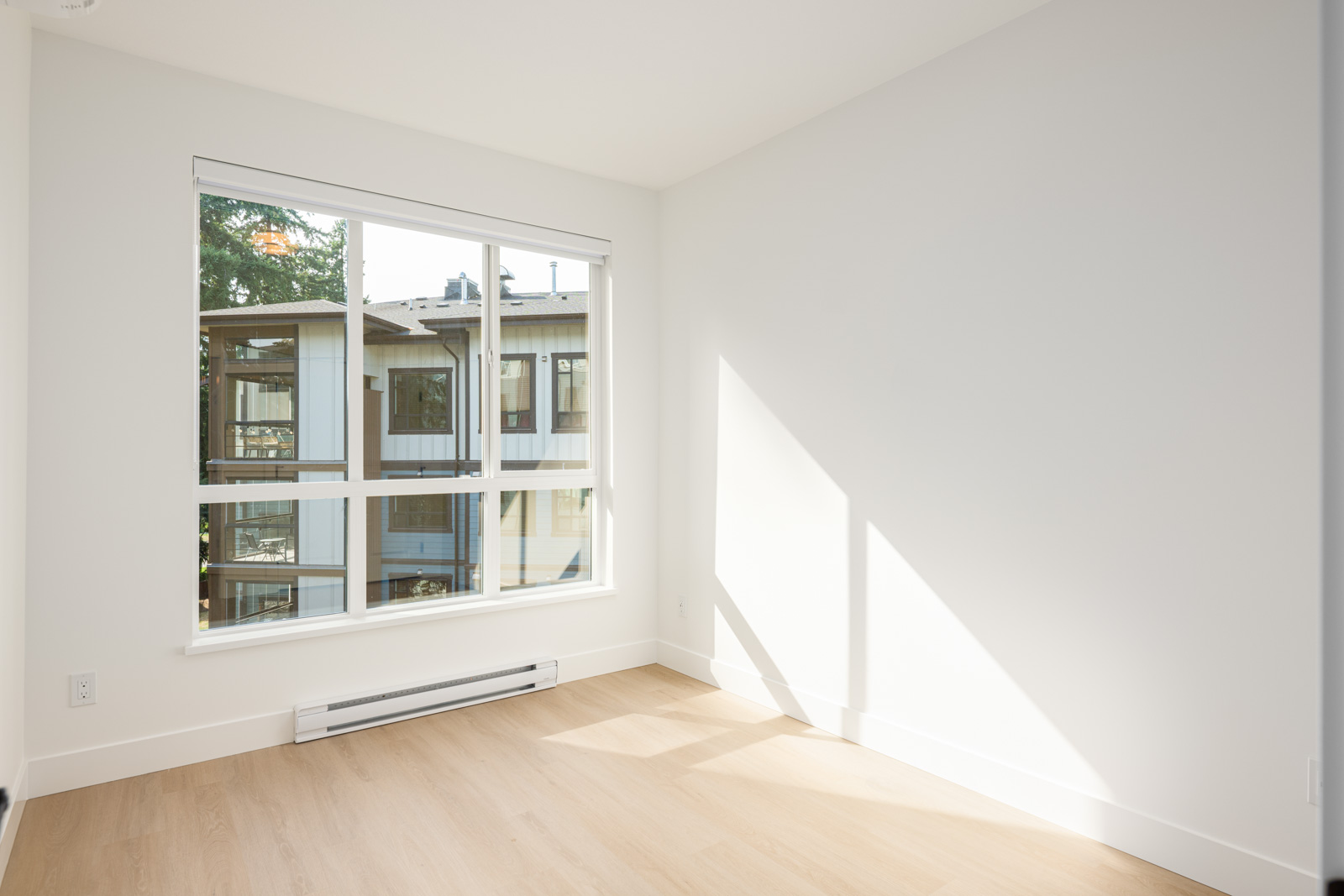 Empty, bright room with light wood flooring, white walls, a large window, and sunlight streaming in. Apartment building visible outside through the window.