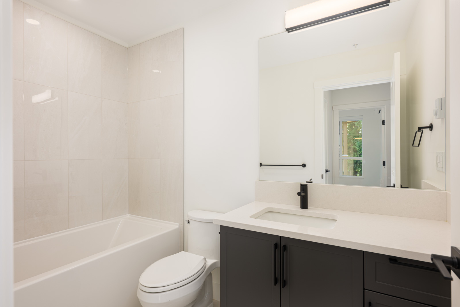 Modern bathroom with a white bathtub and toilet, black vanity with a white countertop, large mirror, and light fixture above the sink.
