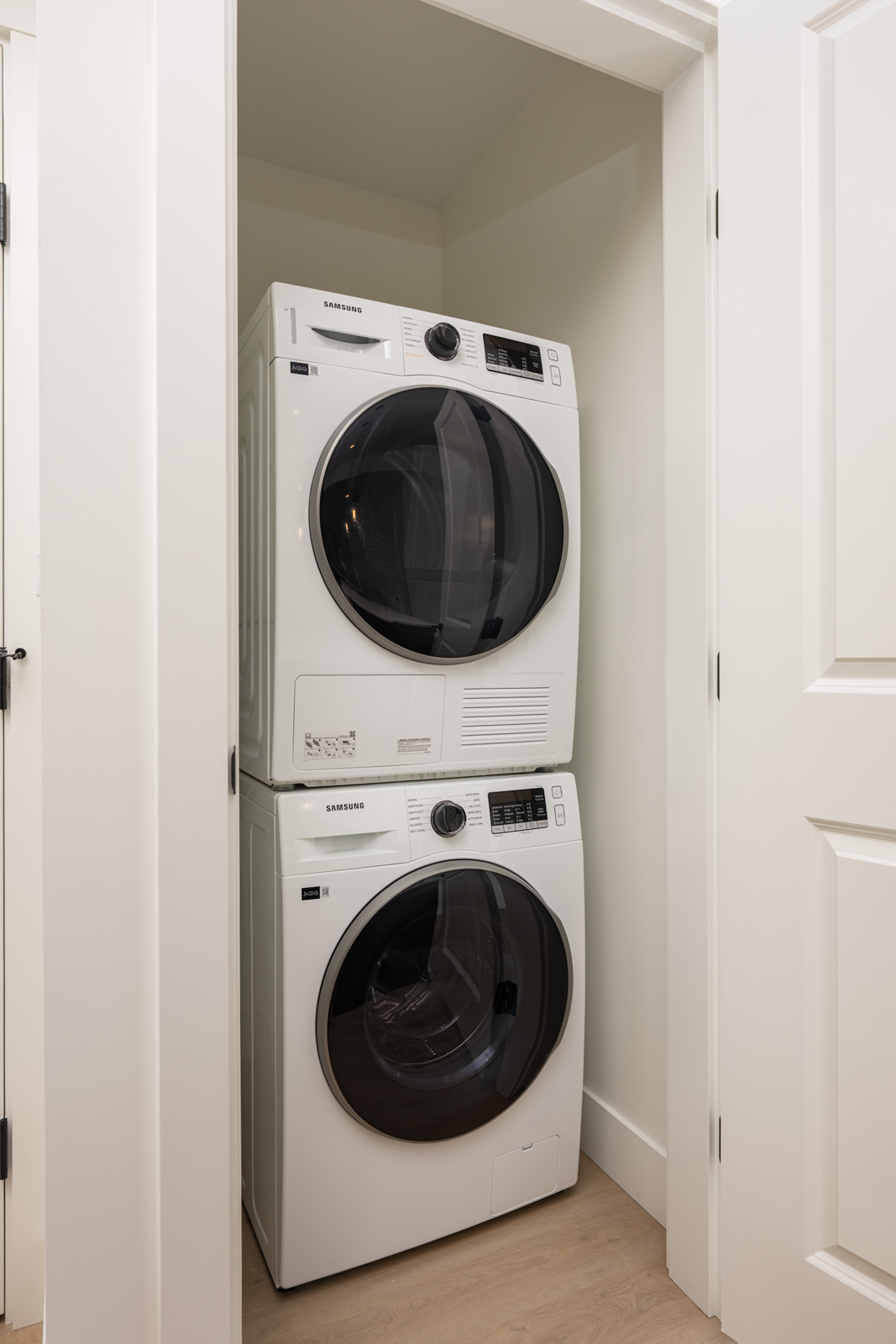 A stacked washer and dryer set is placed in a small, white-walled laundry closet with a light wood floor.